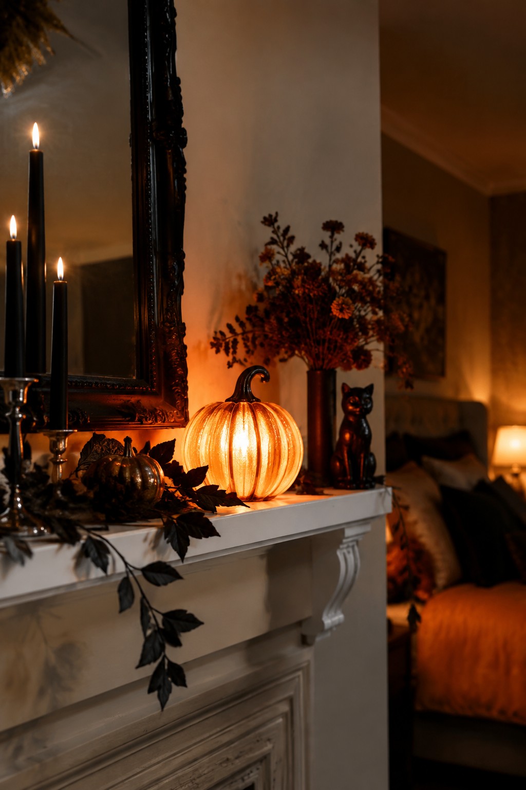 White fireplace mantel with a glowing ribbed pumpkin night light, black taper candles, and dark autumn leaves.