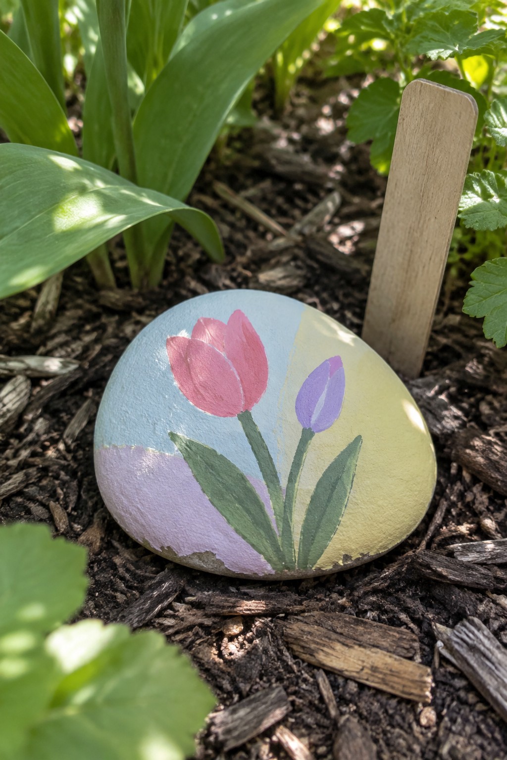 Painted rock with pink and purple tulips among garden plants and soil