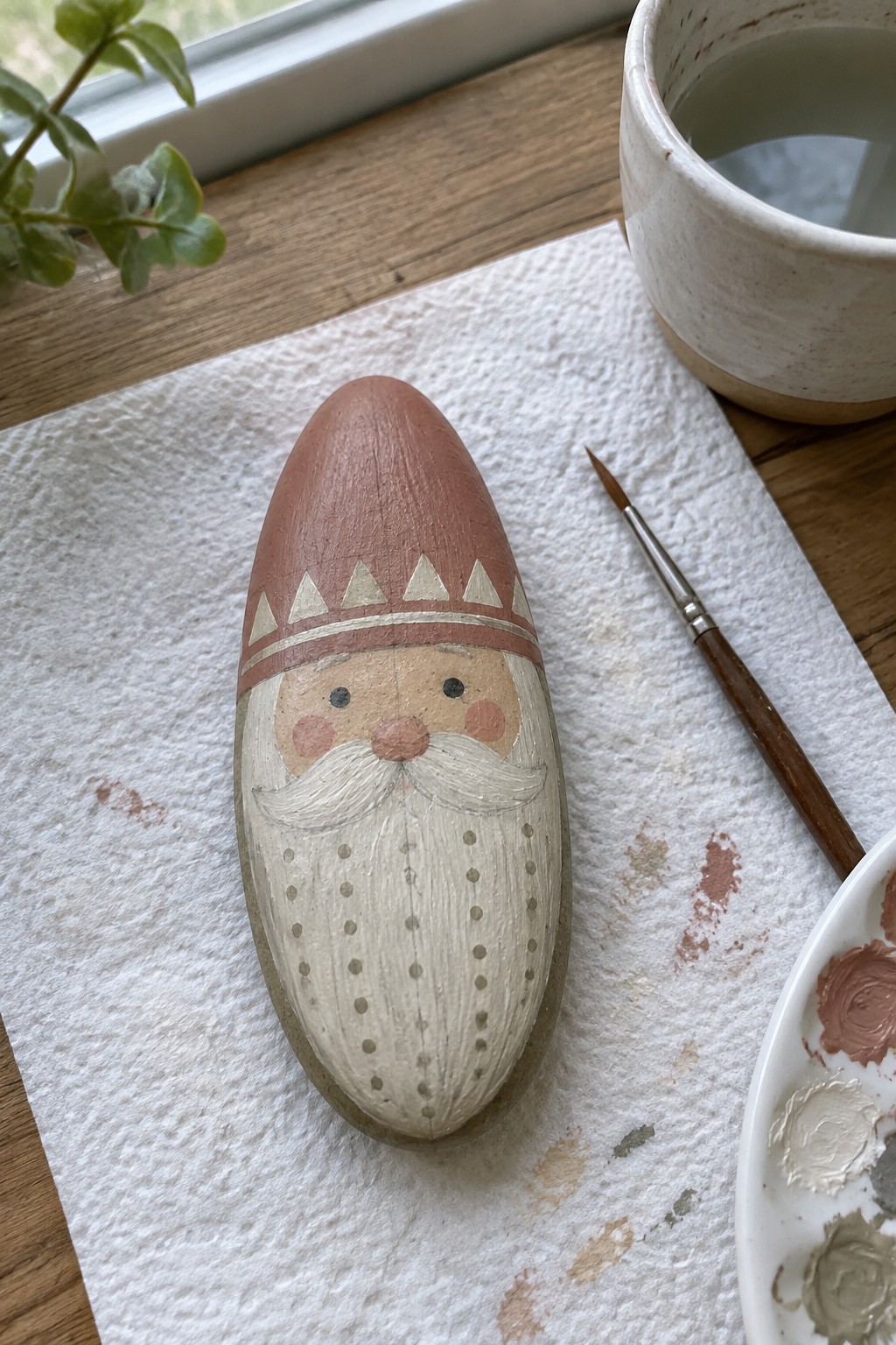 Hand-painted rock gnome with dotted beard and hat beside paintbrush on paper towel.