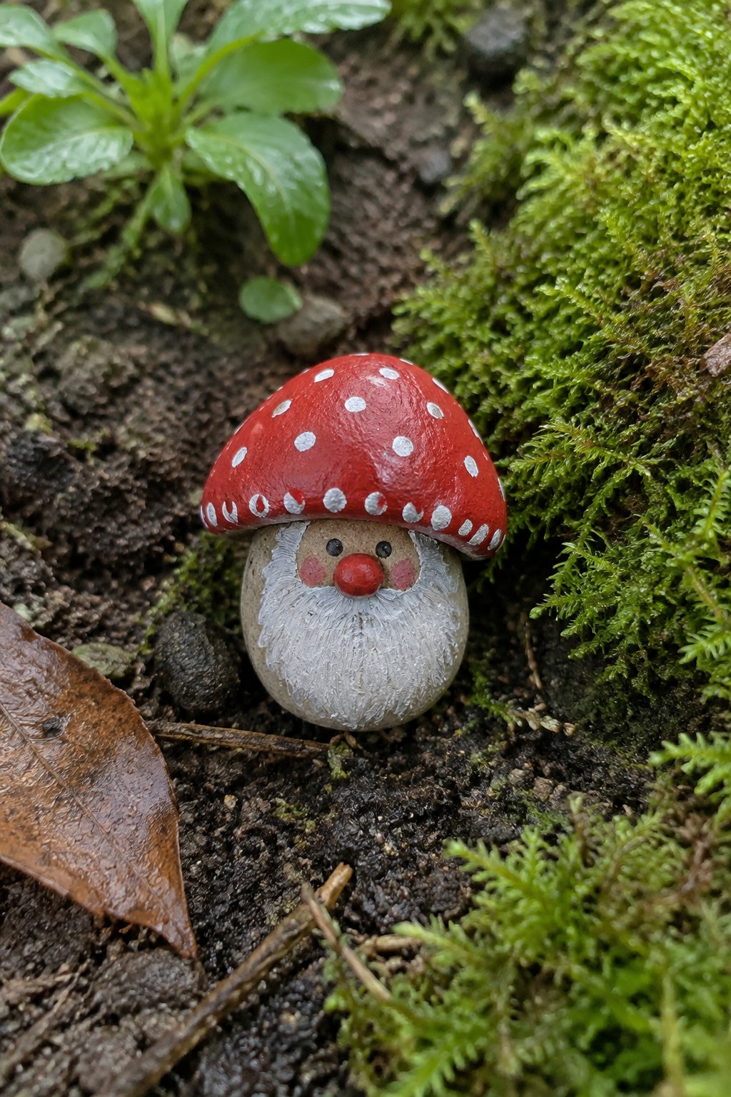 Red mushroom gnome figurine with white polka dots on mossy forest ground.