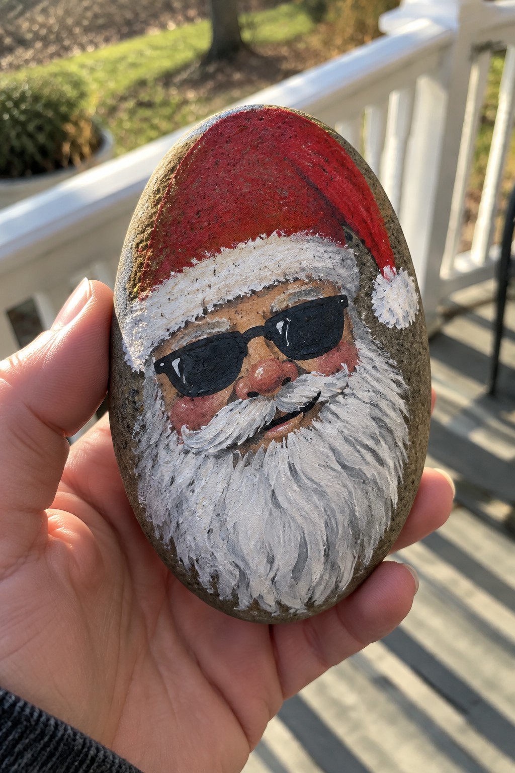 Hand holding rock painted as Santa with sunglasses, red hat, and white beard.