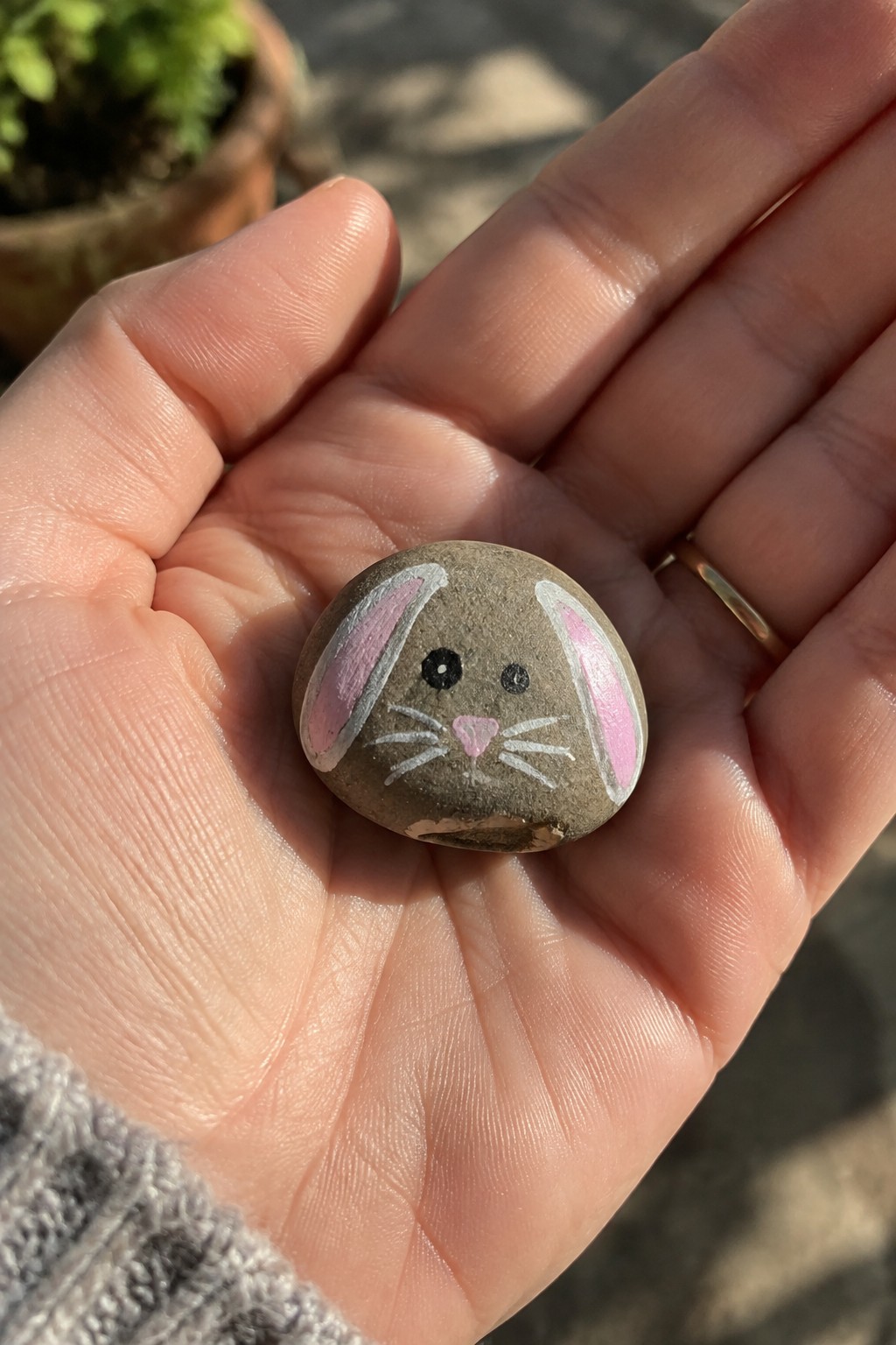 Hand with gold ring holds gray rock painted as bunny with pink ears.