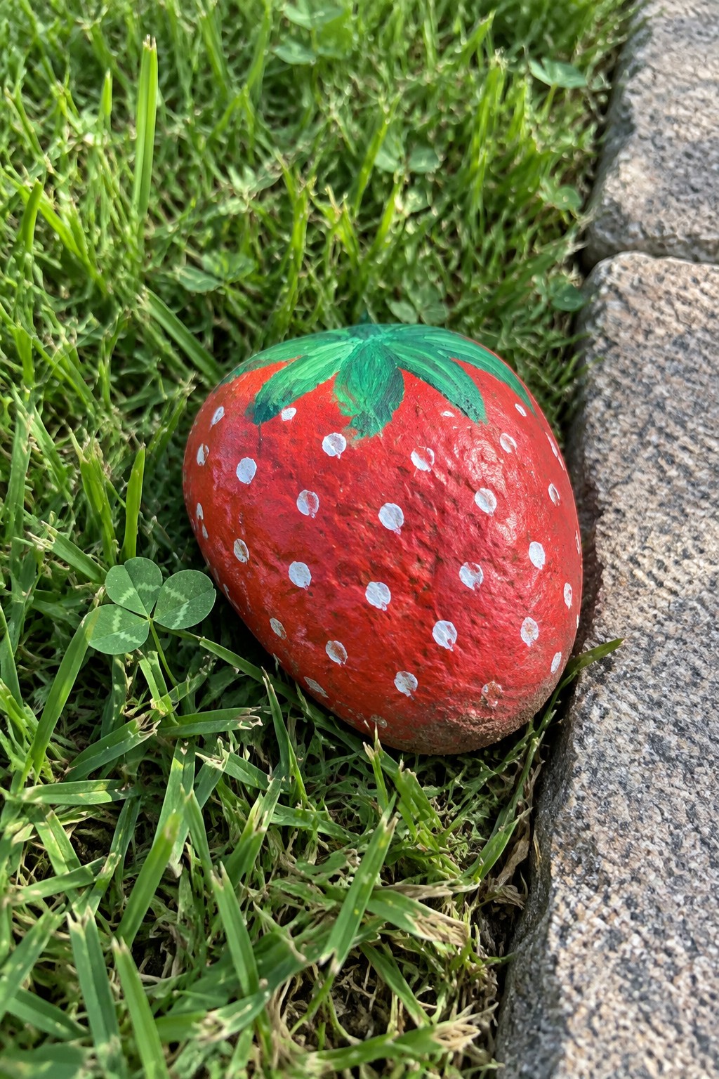 Red strawberry rock with white dots and green leaves on grass by stone edge