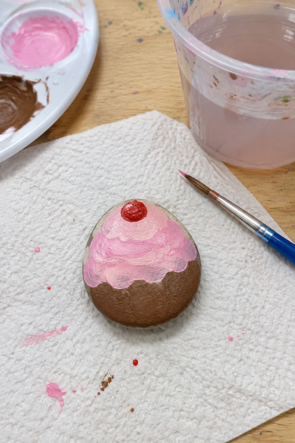Painted stone cupcake with pink frosting, red cherry, on paper towel near paint supplies.
