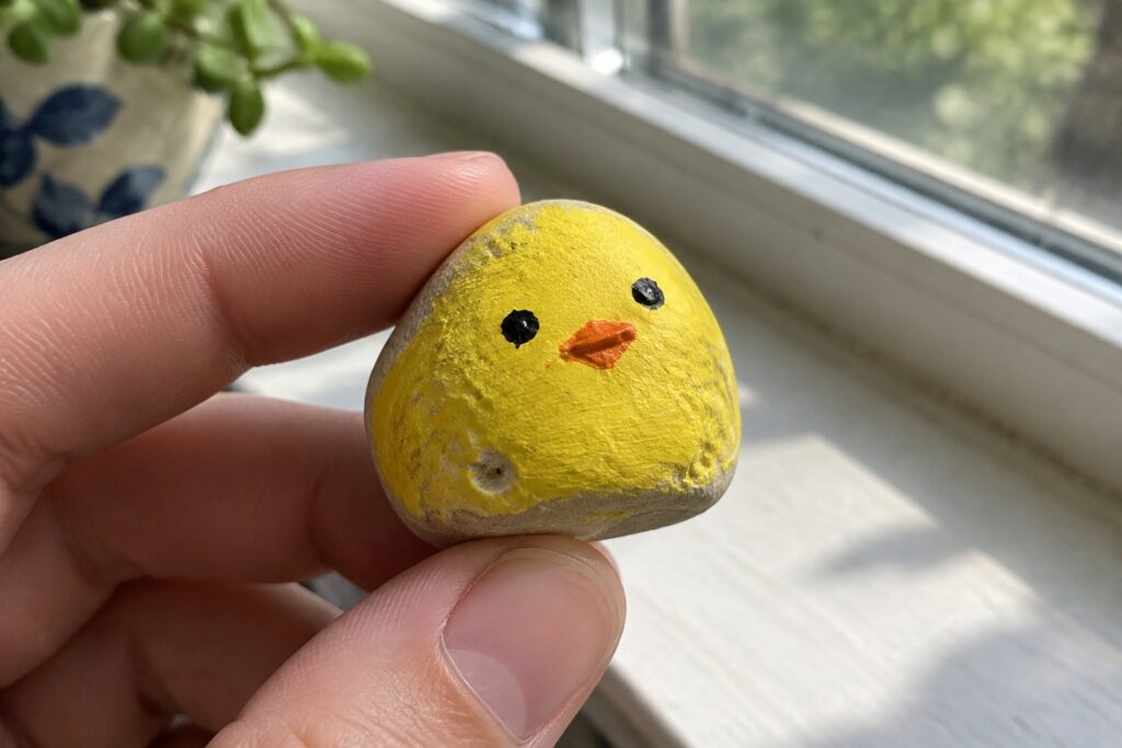21 Creative Small Rock Painting Ideas for Beginners Using Simple Shapes Hand holding small yellow rock painted as chick with black eyes and orange beak