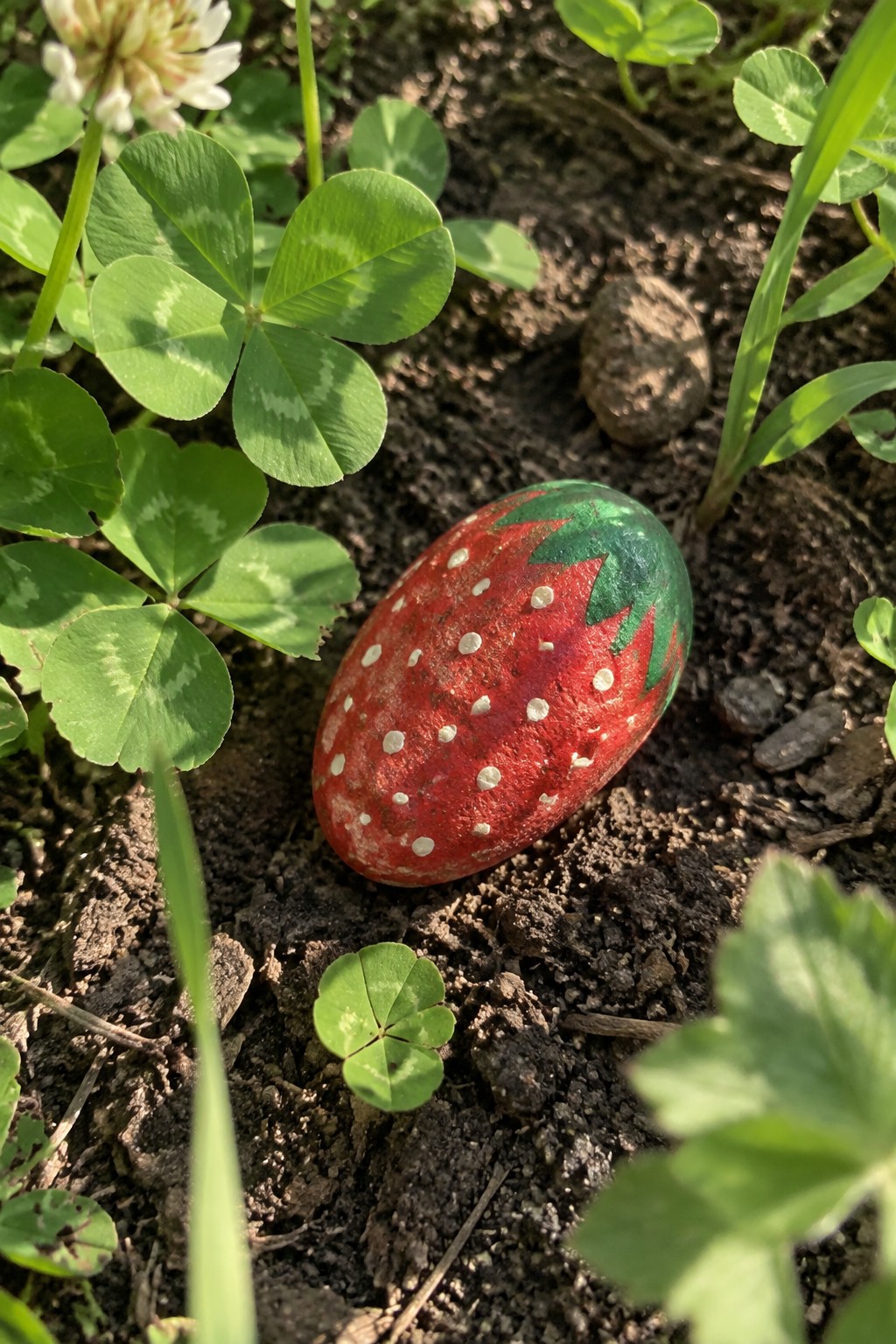 Painted red strawberry rock with white dots and green leaves amid clover on soil