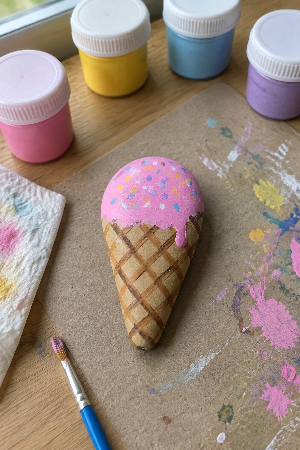 Painted rock ice cream cone with pink frosting and sprinkles amid paint jars and brush