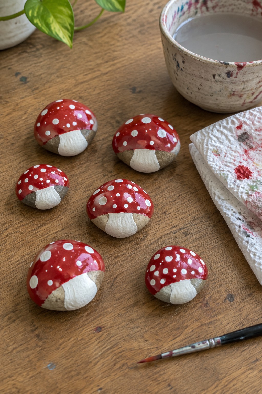 Six red-and-white painted mushroom rocks on wooden table with art supplies.