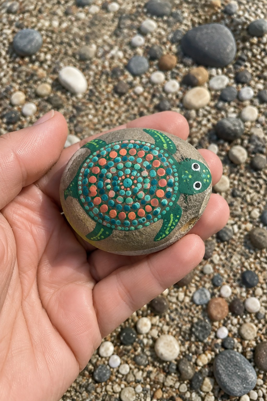 Hand holding a stone painted as a turtle with colorful dotted shell on pebble beach.