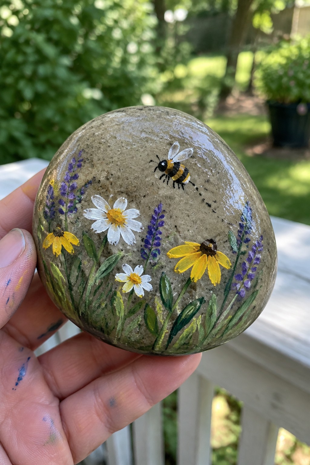 Hand holding glossy painted rock with bee, daisies, and purple flowers.