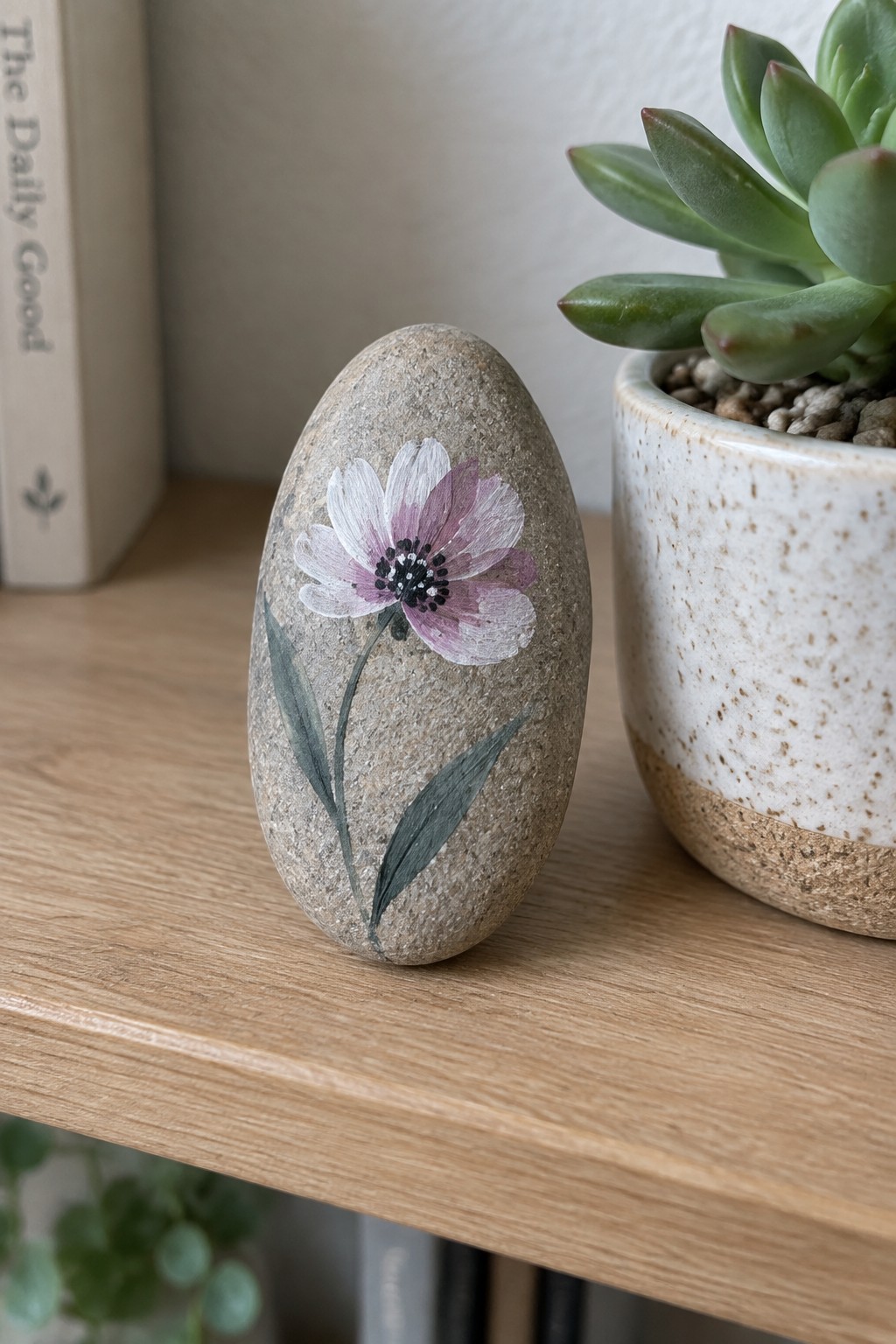 Oval stone painted with purple flower beside potted succulent on wooden shelf
