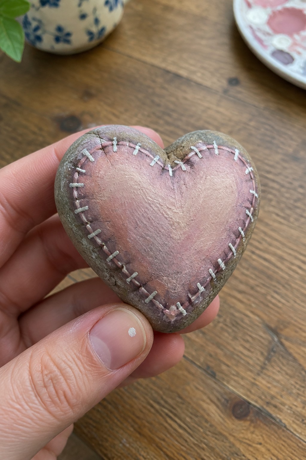 Hand holding pink heart-shaped stone mended with white stitches on wood.