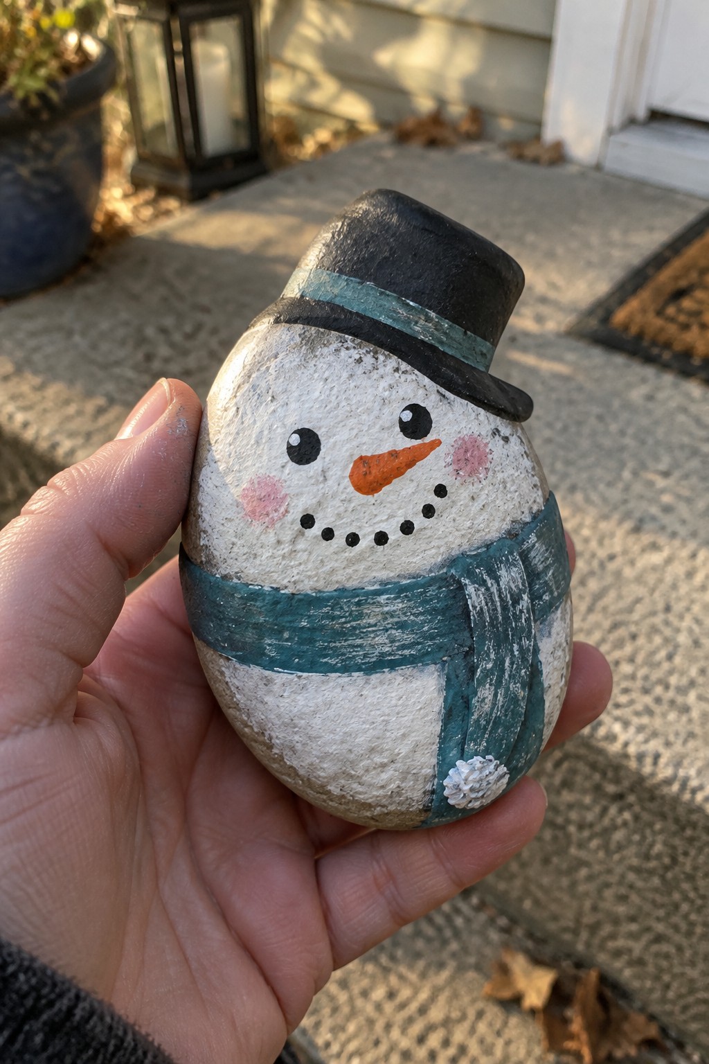 A hand holds a painted oval rock snowman featuring a black top hat with teal band, teal scarf, and orange nose.