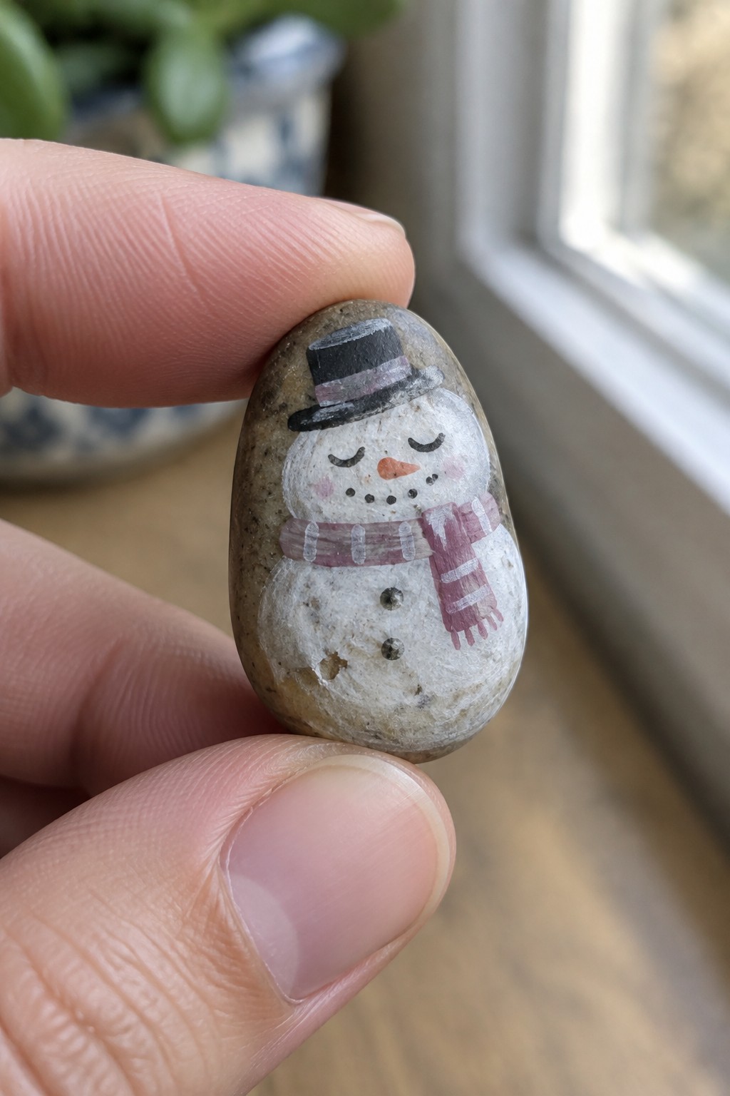 Hand holding pebble painted as snowman with top hat and striped scarf