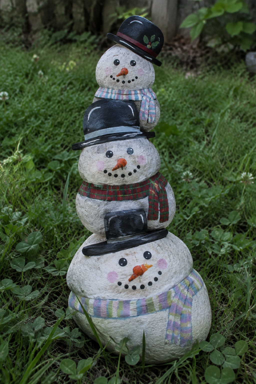 Three stacked painted rocks as snowmen with hats and scarves in grass.