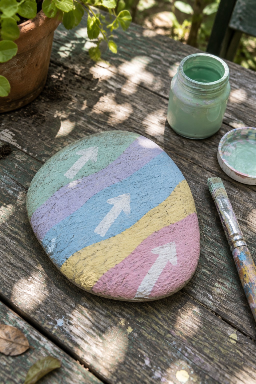 Painted rock with pastel stripes and white arrows beside green paint jar and brush