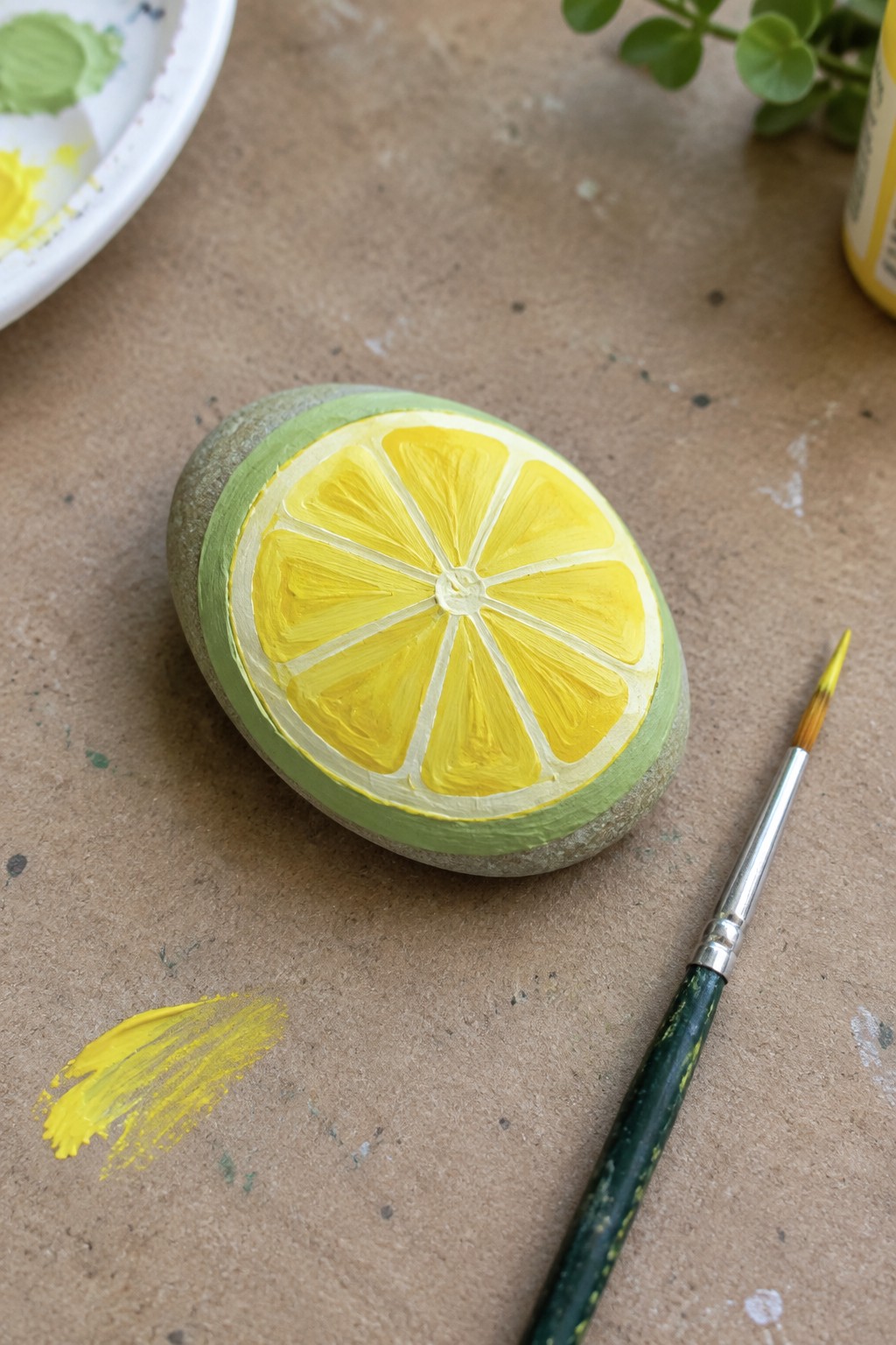 Painted rock as lemon slice with yellow segments, green rind, beside brush and paint smear.