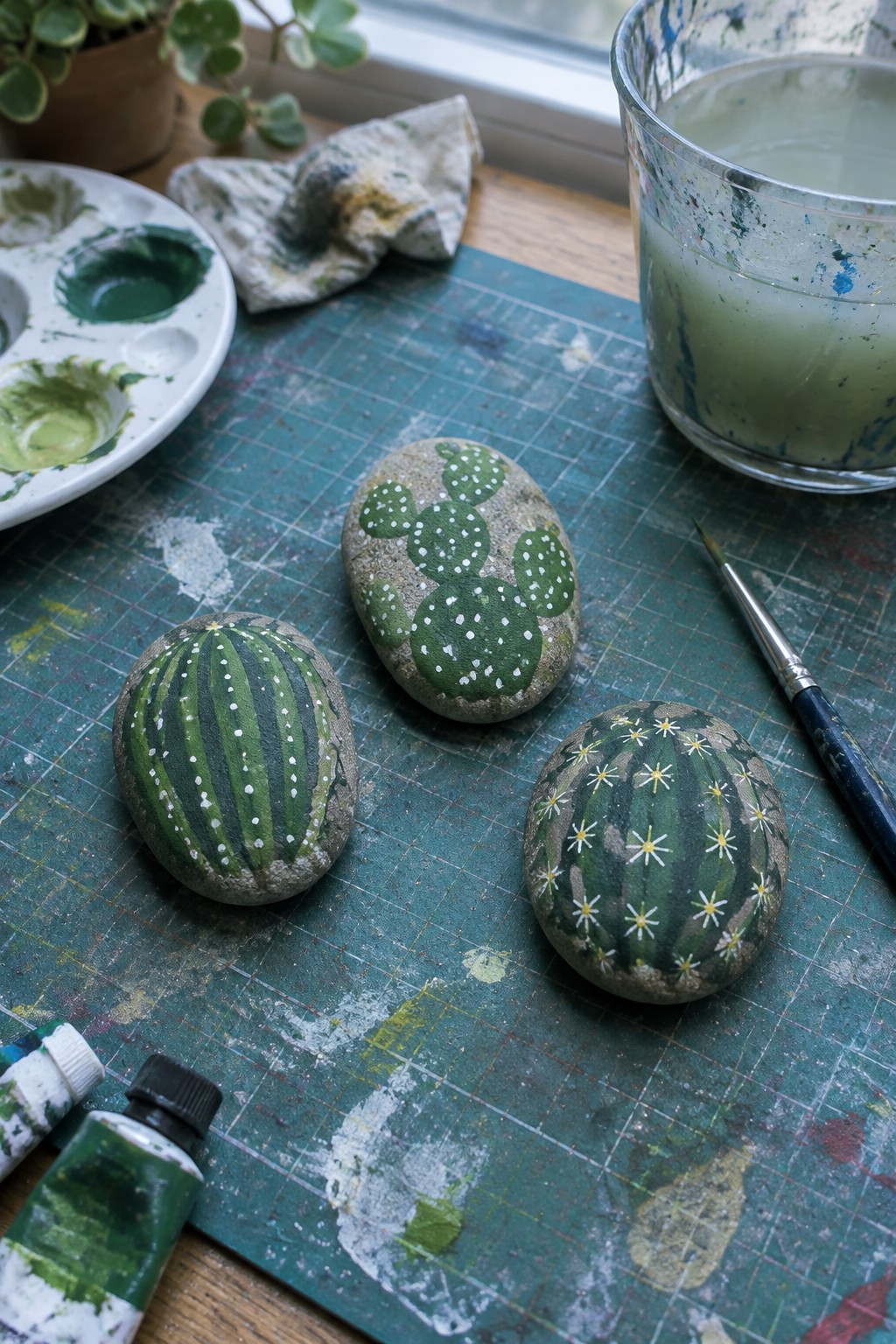 Painted rocks with cactus and watermelon designs on green mat amid paints and brush