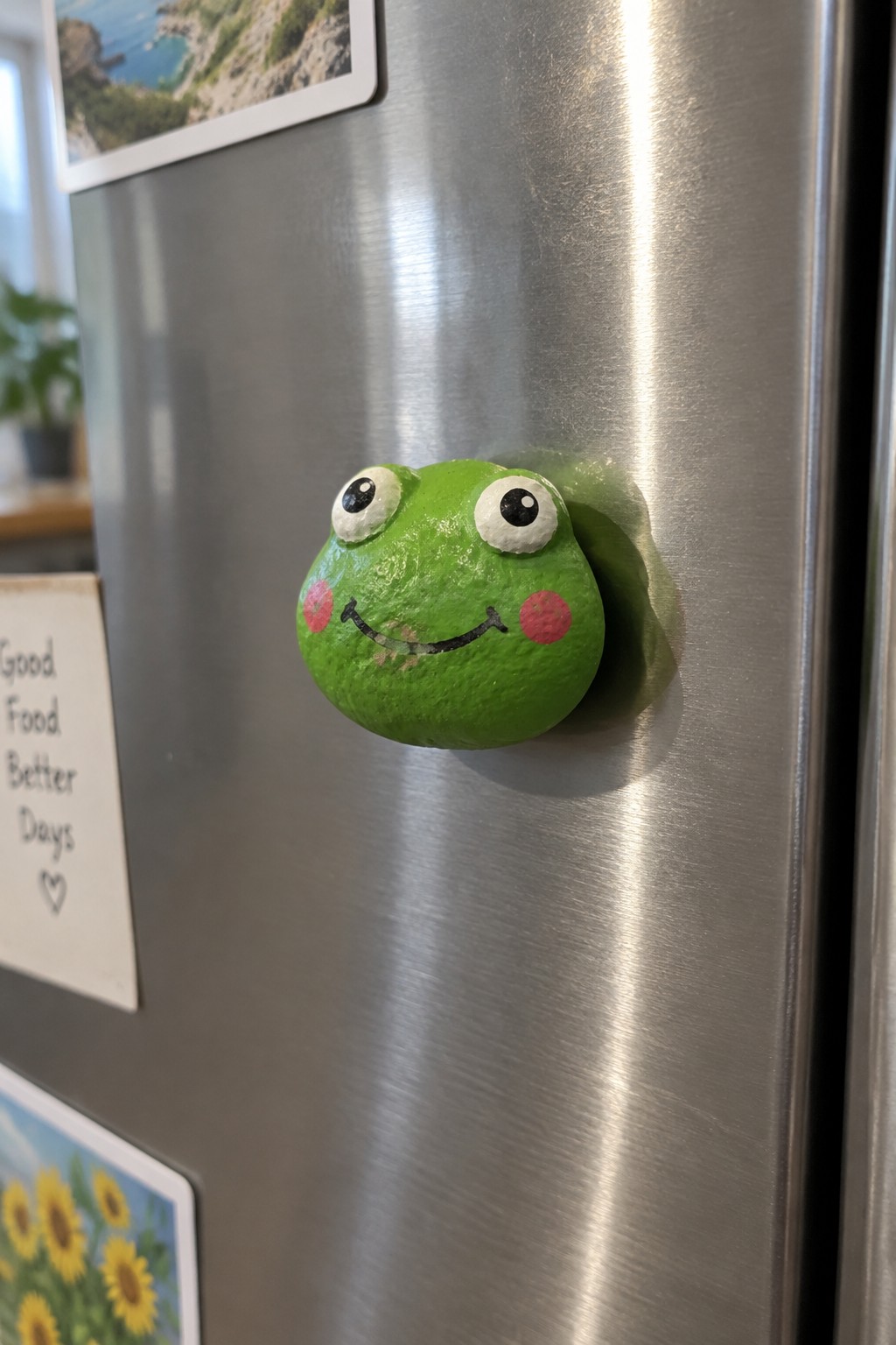 Green frog magnet with large eyes and pink cheeks on stainless steel fridge