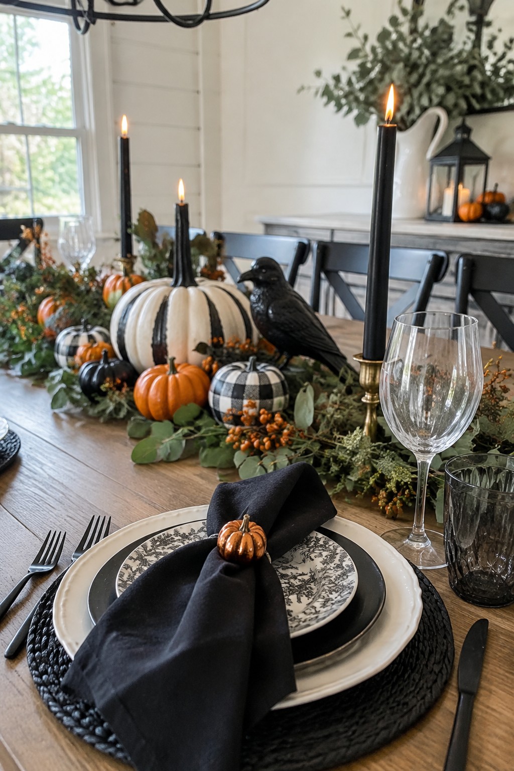 Dining table with a long centerpiece of pumpkins, greenery, black candles, and a black crow, plus black napkins and stacked plates at each setting.