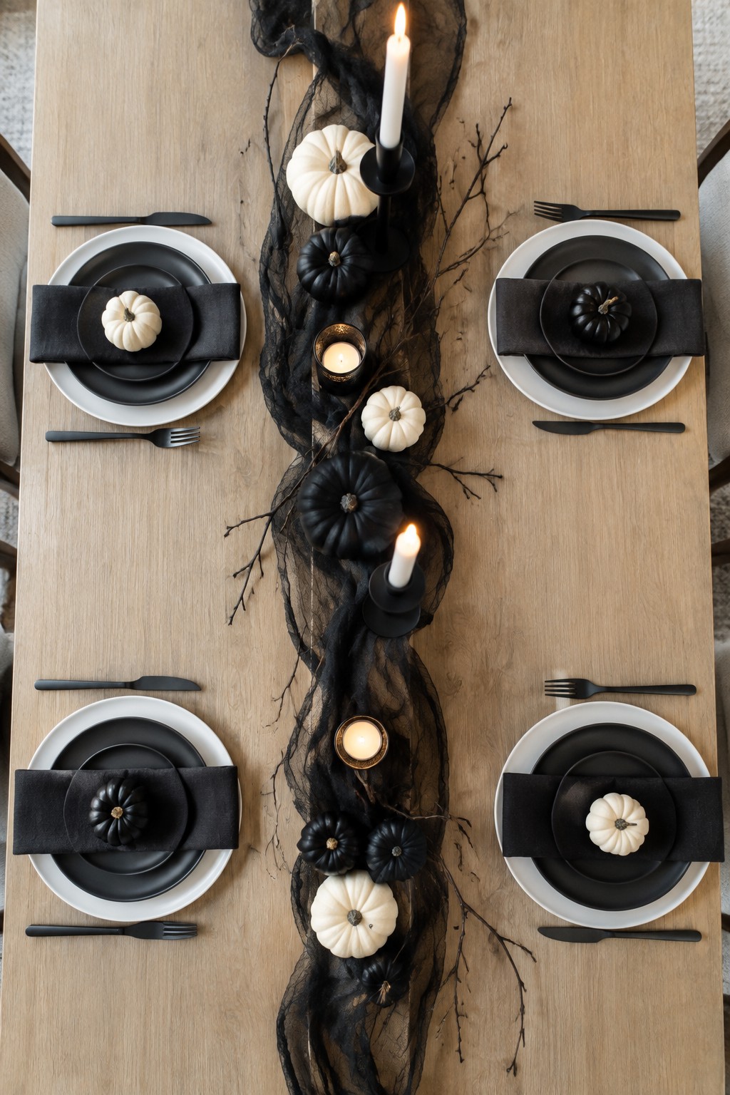 A wooden dining table set for four with black and white plates, black napkins, and a long centerpiece of black sheer fabric, black branches, black and white pumpkins, and lit candles.