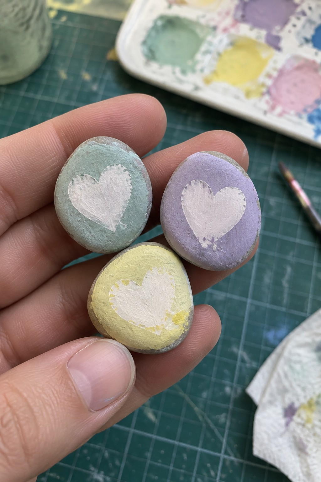 Hand holding three pastel stones painted with white hearts on green mat