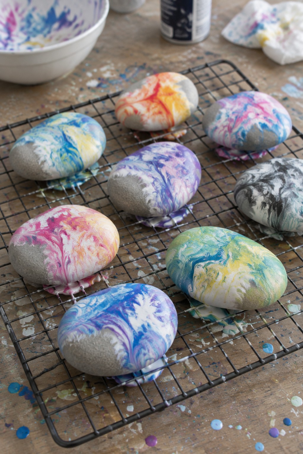 Colorful marbled painted rocks drying on a wire cooling rack with art supplies nearby