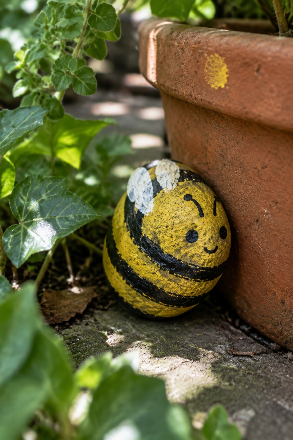 Smiling bee painted on rock leaning against weathered terracotta pot with plants