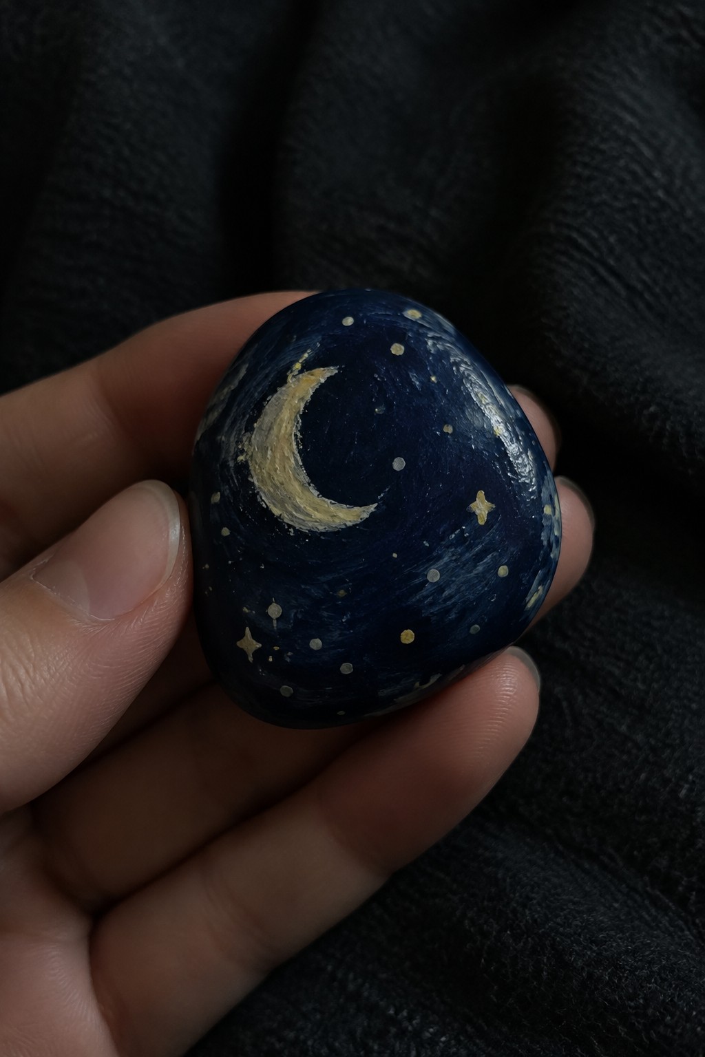 Hand holding smooth dark blue stone painted with yellow crescent moon and stars.