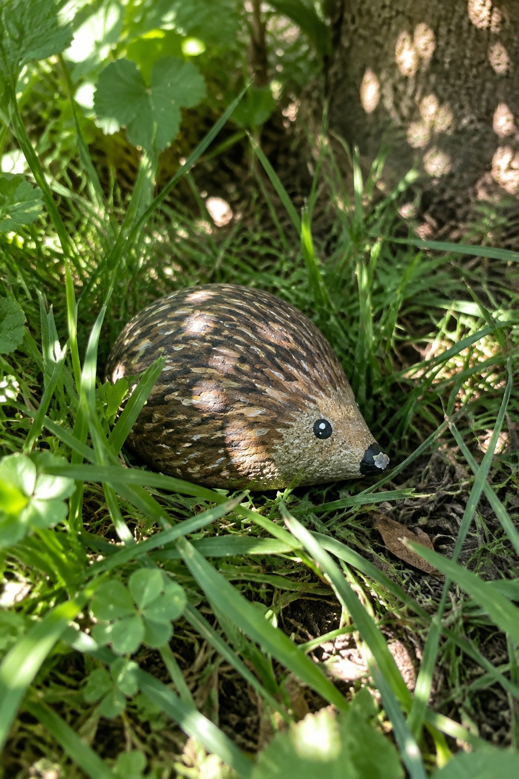 Painted brown hedgehog rock nestled in green grass near tree trunk.