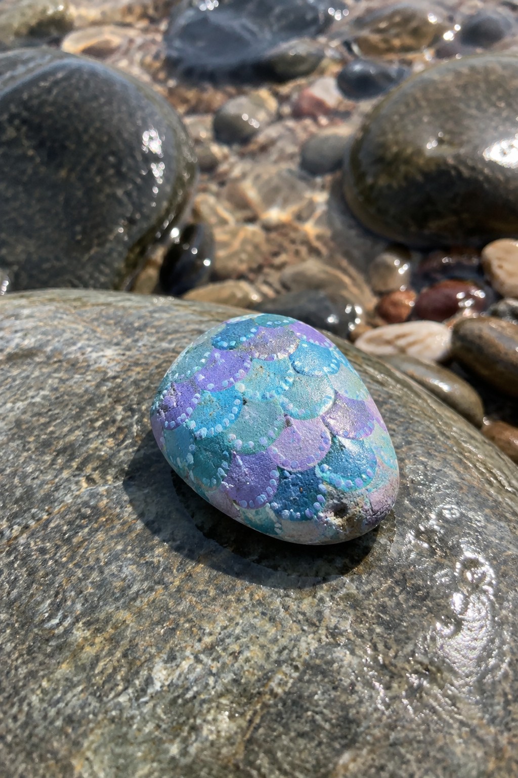 Colorful painted rock with blue-purple mermaid scales on wet gray boulder by water
