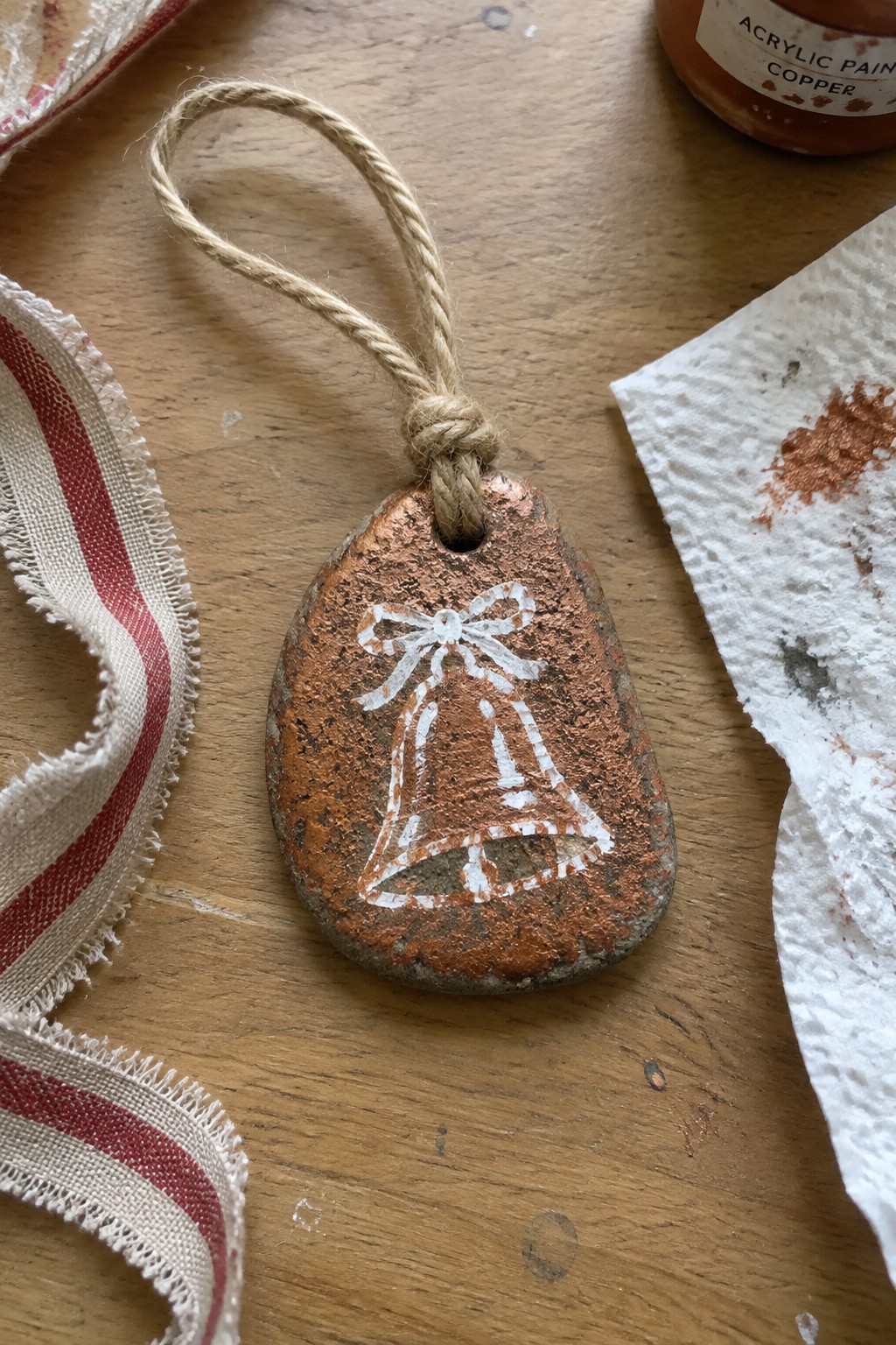 Copper-painted stone ornament with white bell and bow hangs from twine on wood.