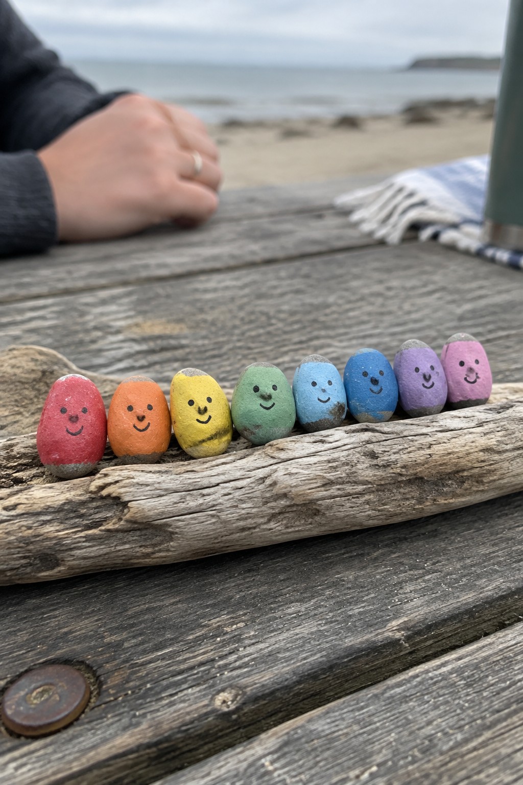 Rainbow smiley rocks lined on driftwood atop beachside wooden table with hand nearby.
