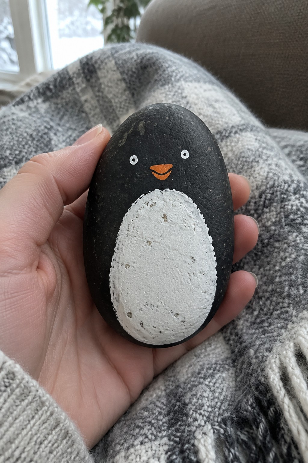 A hand holds a black oval rock painted with a white belly, orange beak, and small white eyes to resemble a penguin.