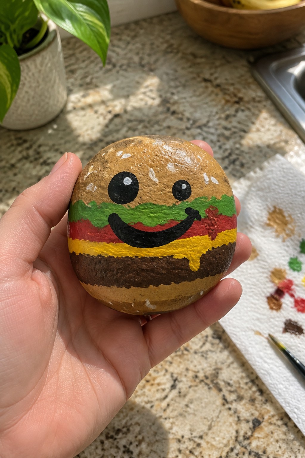 Hand holding a smiling hamburger rock painted with layered colors on kitchen counter.