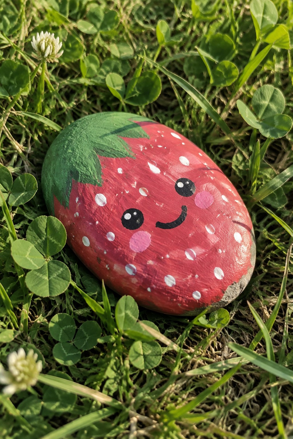 Smiling strawberry painted rock on lush green grass with clover leaves