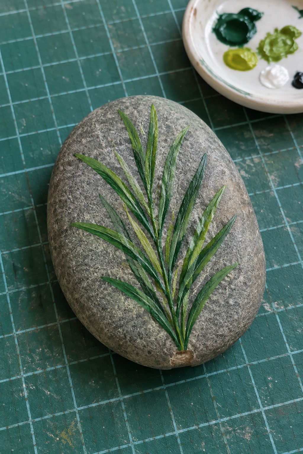 Oval gray rock with painted green herb sprig and paint palette visible in background.