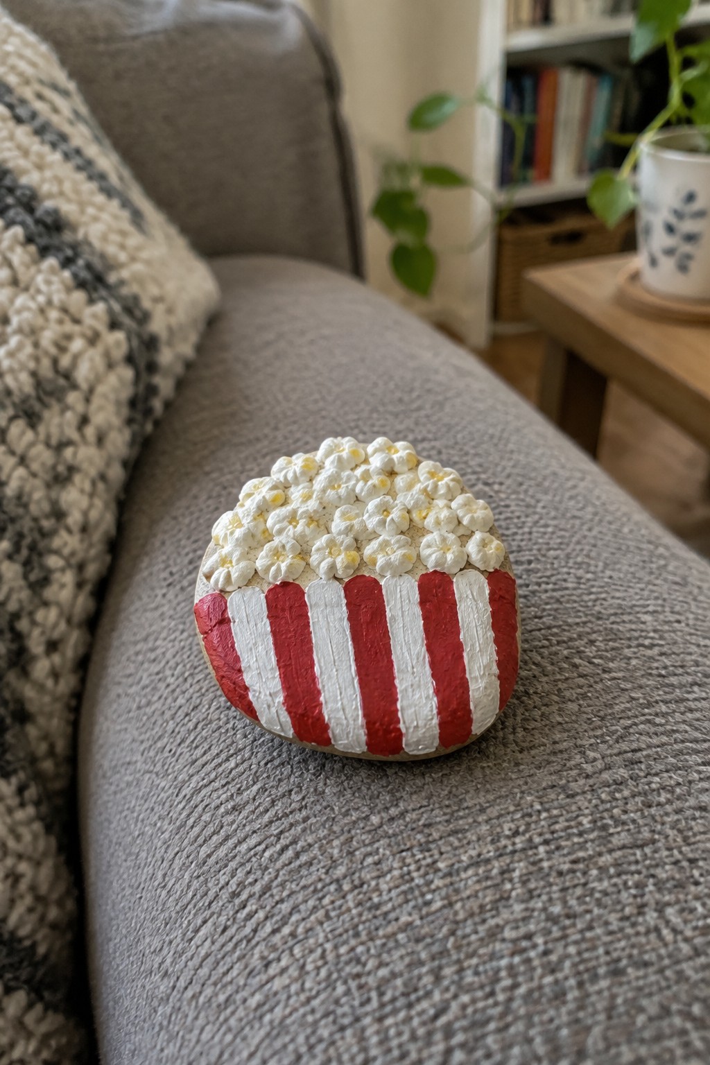 Painted rock shaped like popcorn in red-and-white striped bucket on gray couch