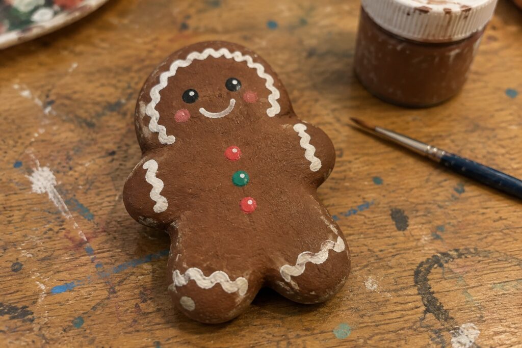 Painted gingerbread man rock with white icing and green buttons beside paint jar.