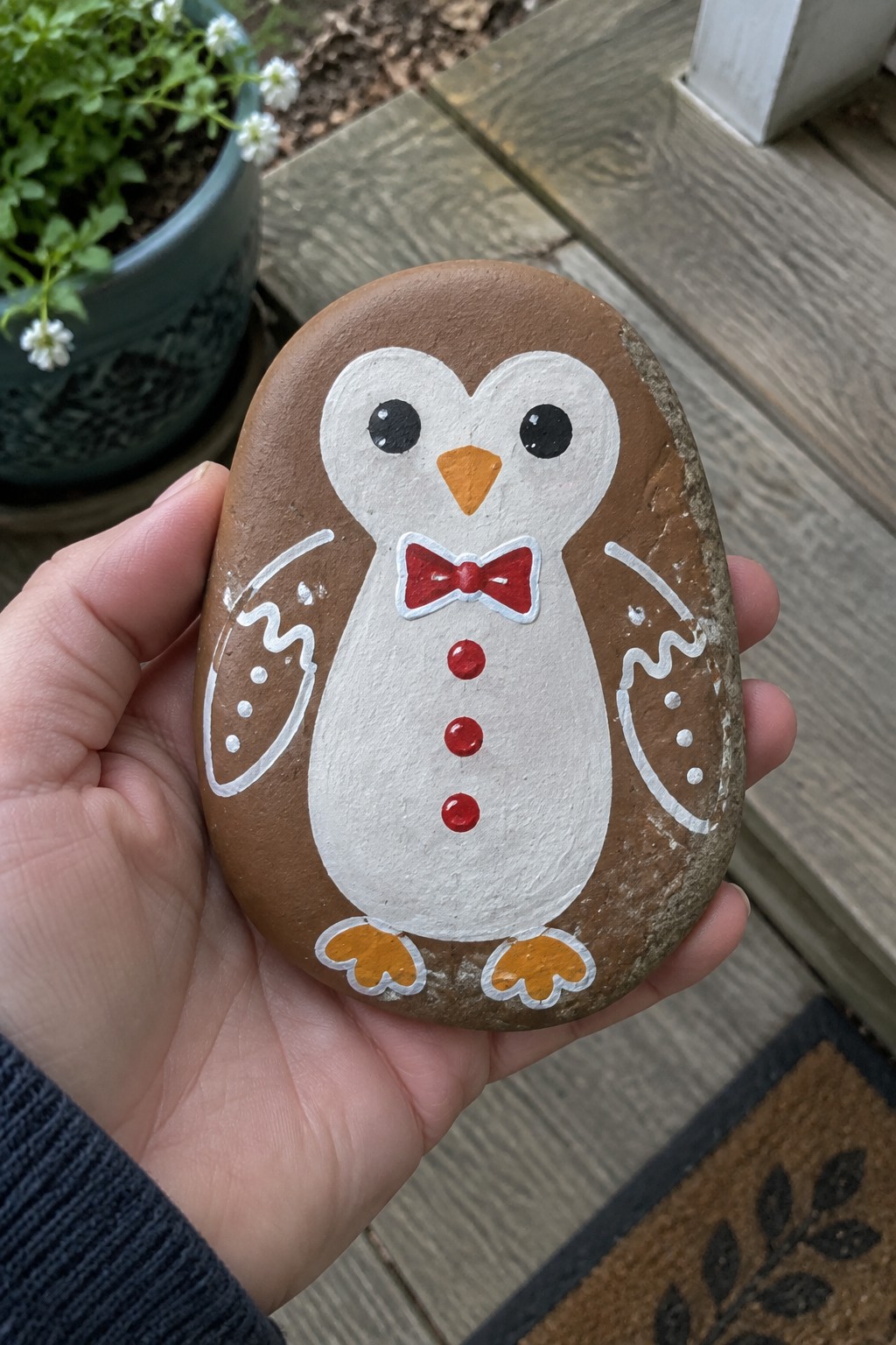 Hand holding oval rock painted as penguin with red bow tie and buttons