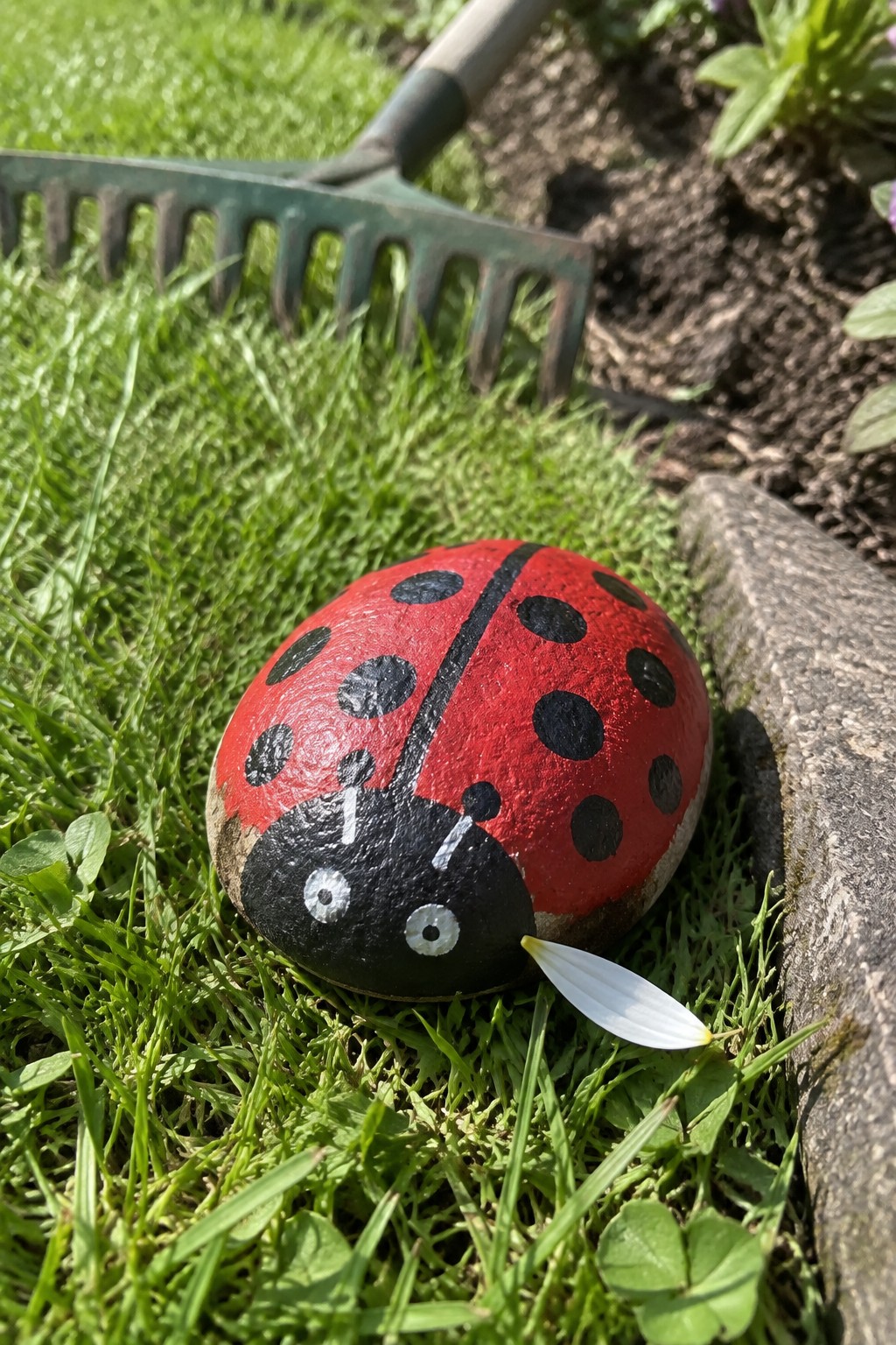 Red ladybug painted on rock in grass near garden rake.