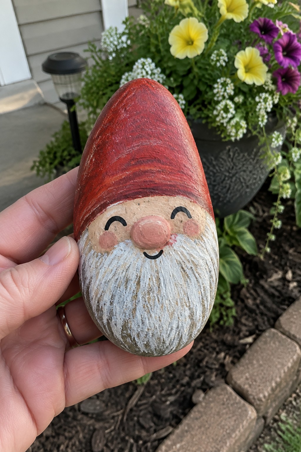 Hand holding painted rock gnome with red hat, white beard, and rosy cheeks in garden