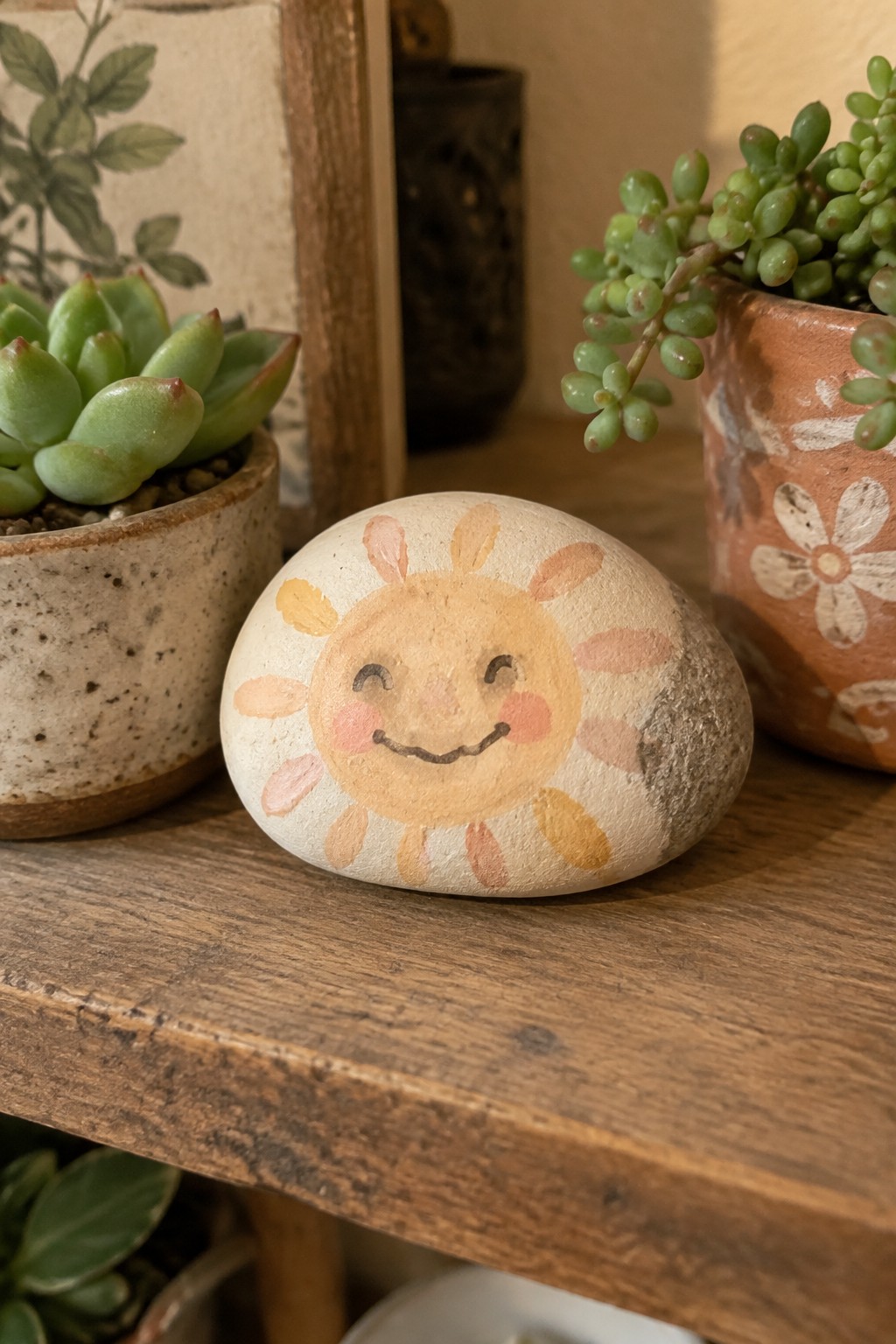Painted smiling sun rock on wooden shelf between potted succulents.