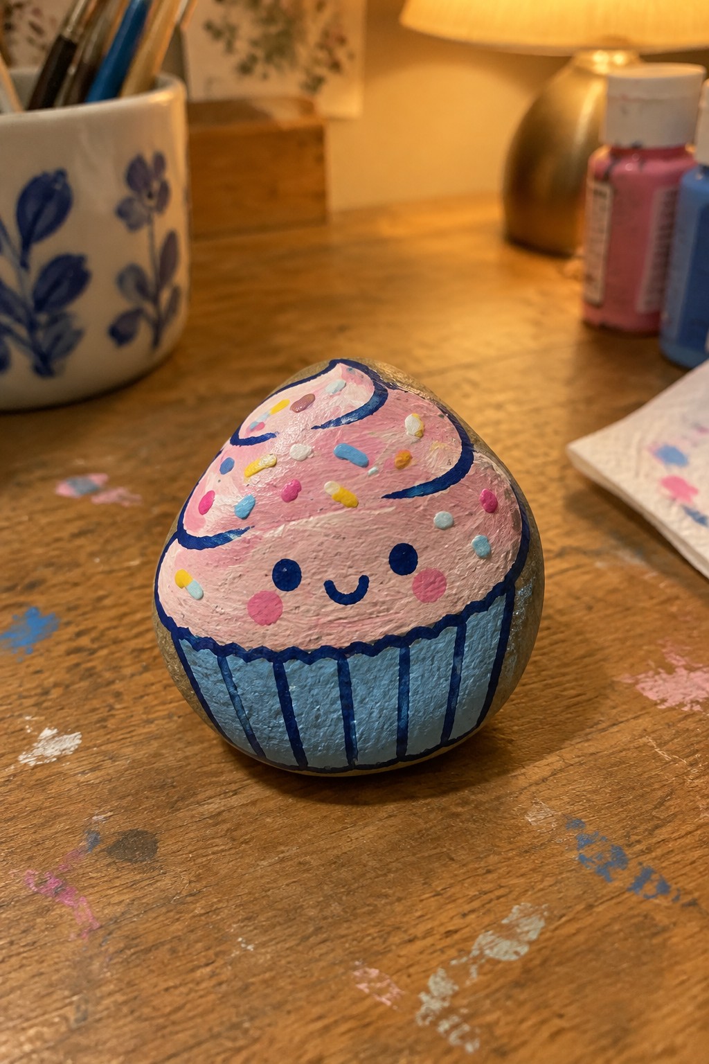 Painted rock as pink cupcake with blue liner, sprinkles, and smiling face on wood table.