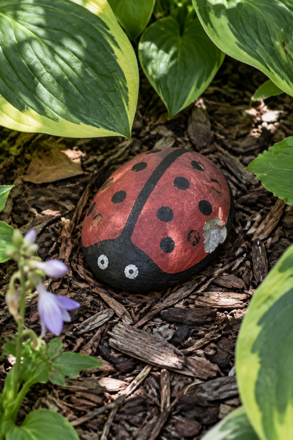 A red ladybug with black spots and a black head painted on a rounded rock in garden mulch.