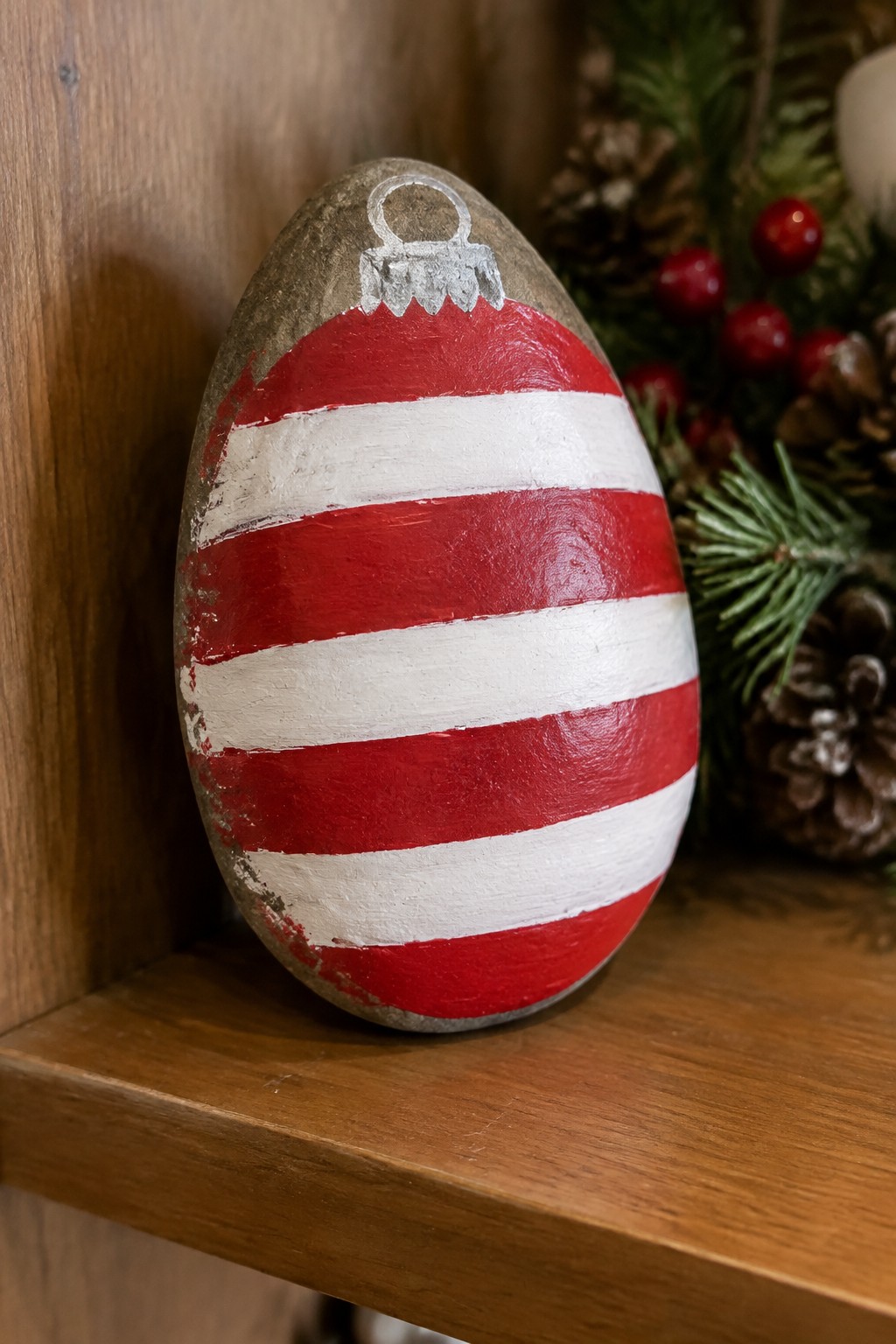Red and white striped painted stone Christmas ornament on wooden shelf with pine branches