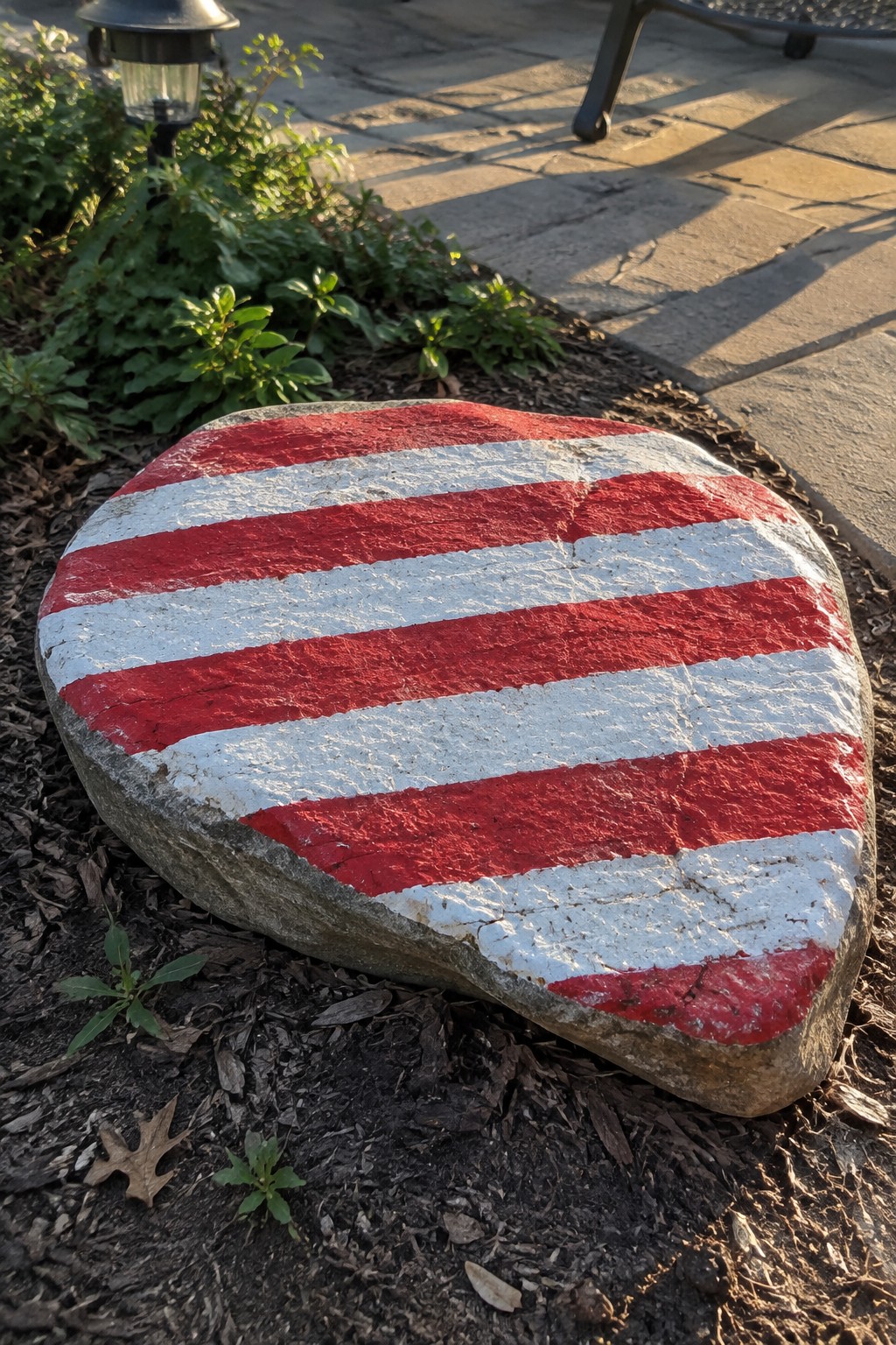 Large rock painted with thick red and white horizontal stripes on garden mulch.