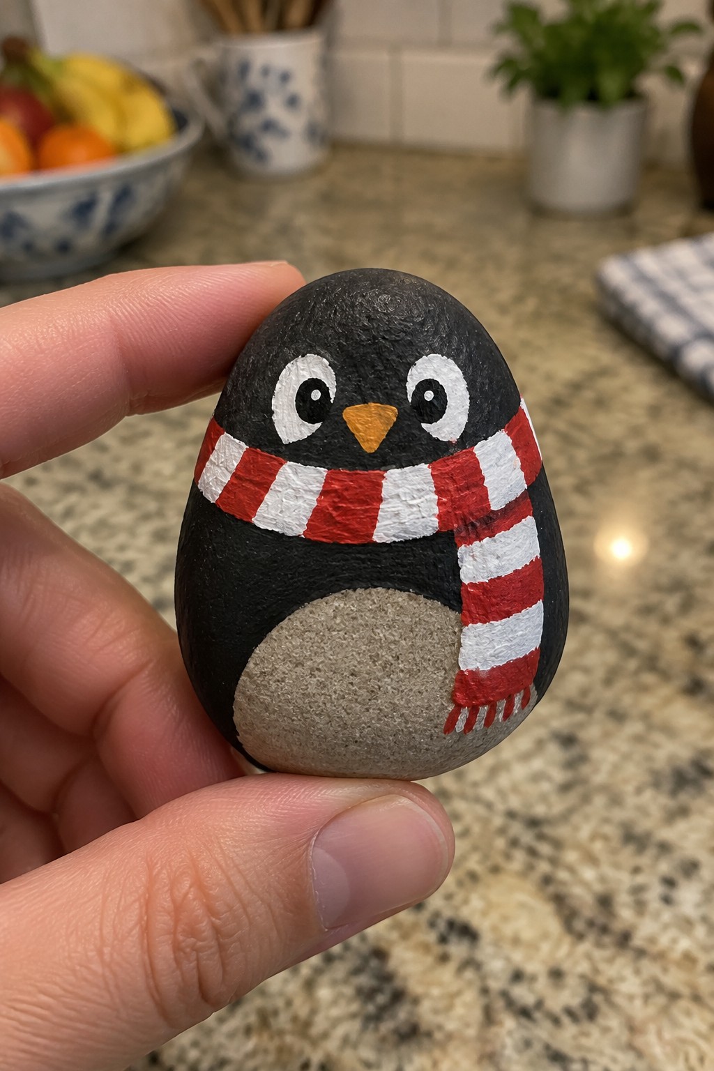 Hand holding black painted rock penguin with red striped scarf and orange beak