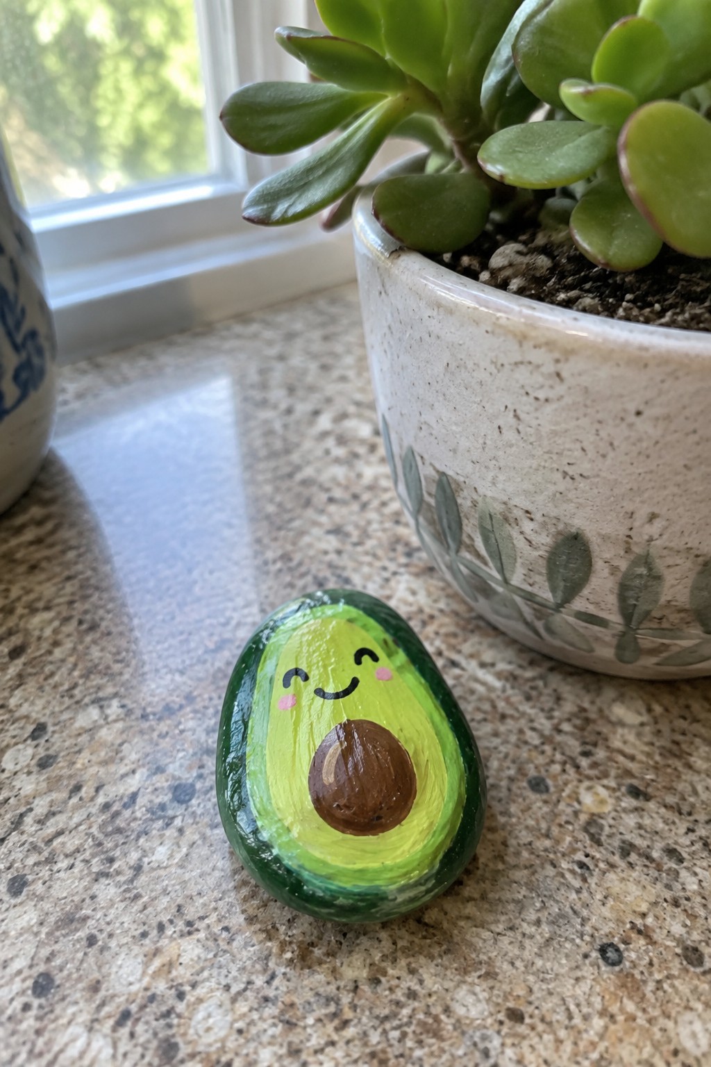 Smiling avocado rock art on granite counter next to leafy succulent in ceramic pot