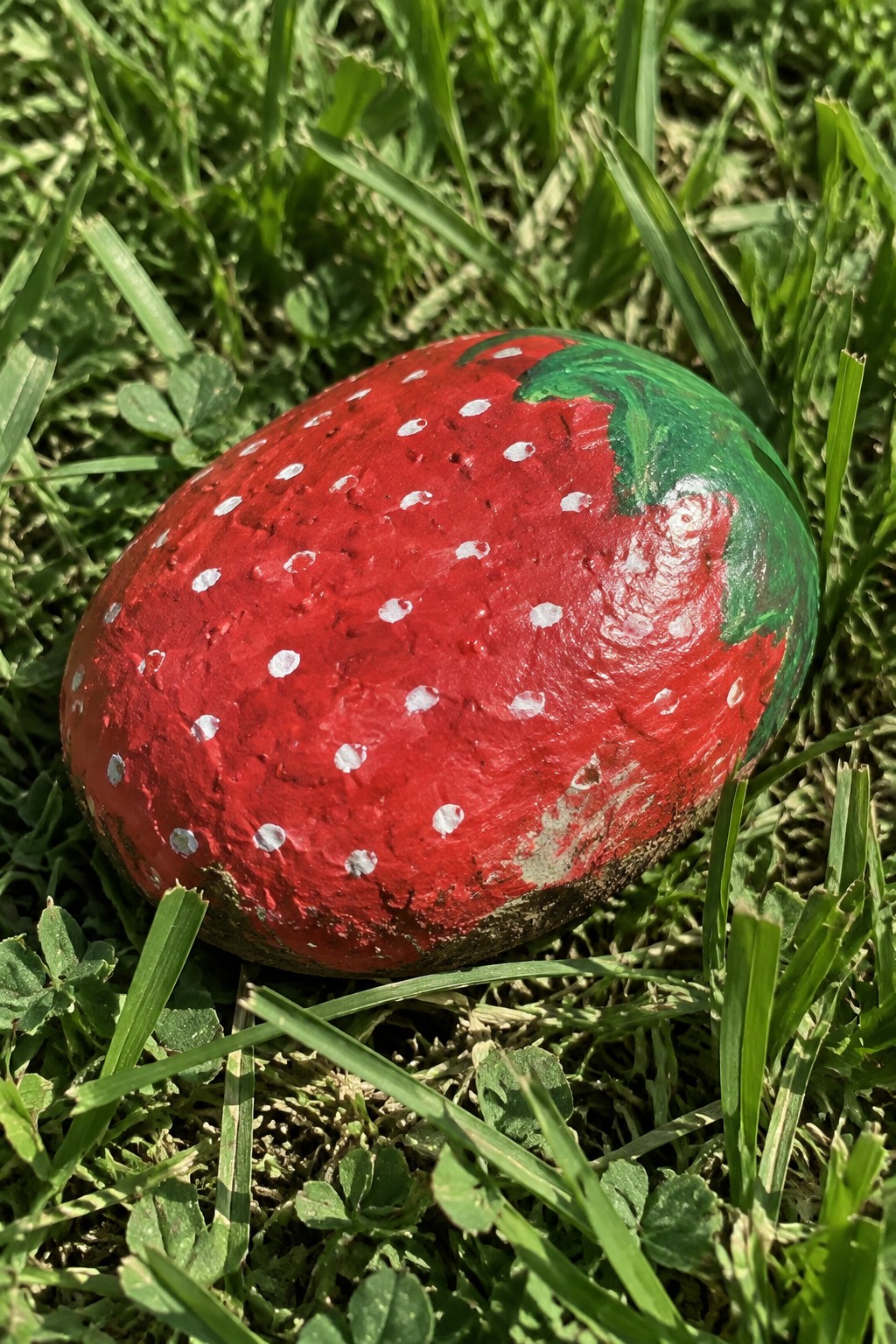 Red strawberry-painted rock with white dots and green leaves on grass